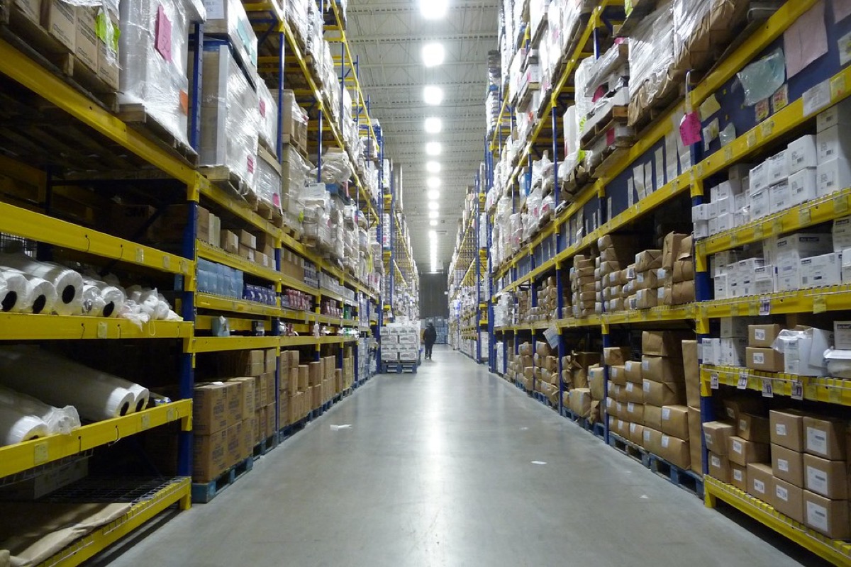 A long warehouse aisle lined with tall yellow and blue pallet racking stacked with boxed and wrapped restaurant supplies.