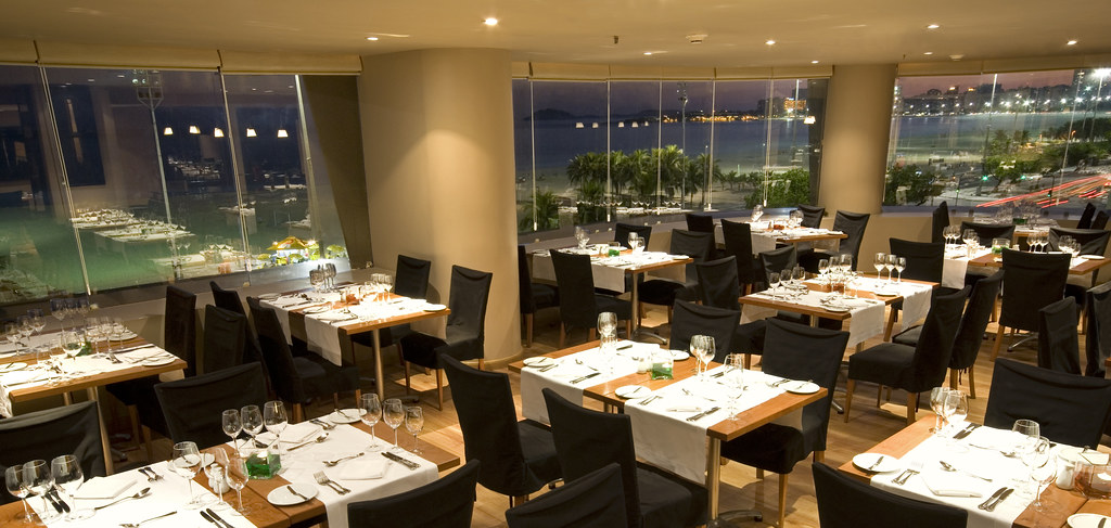 An empty restaurant dining room at dusk with tables set with white linen, glasses and cutlery, city views through the windows