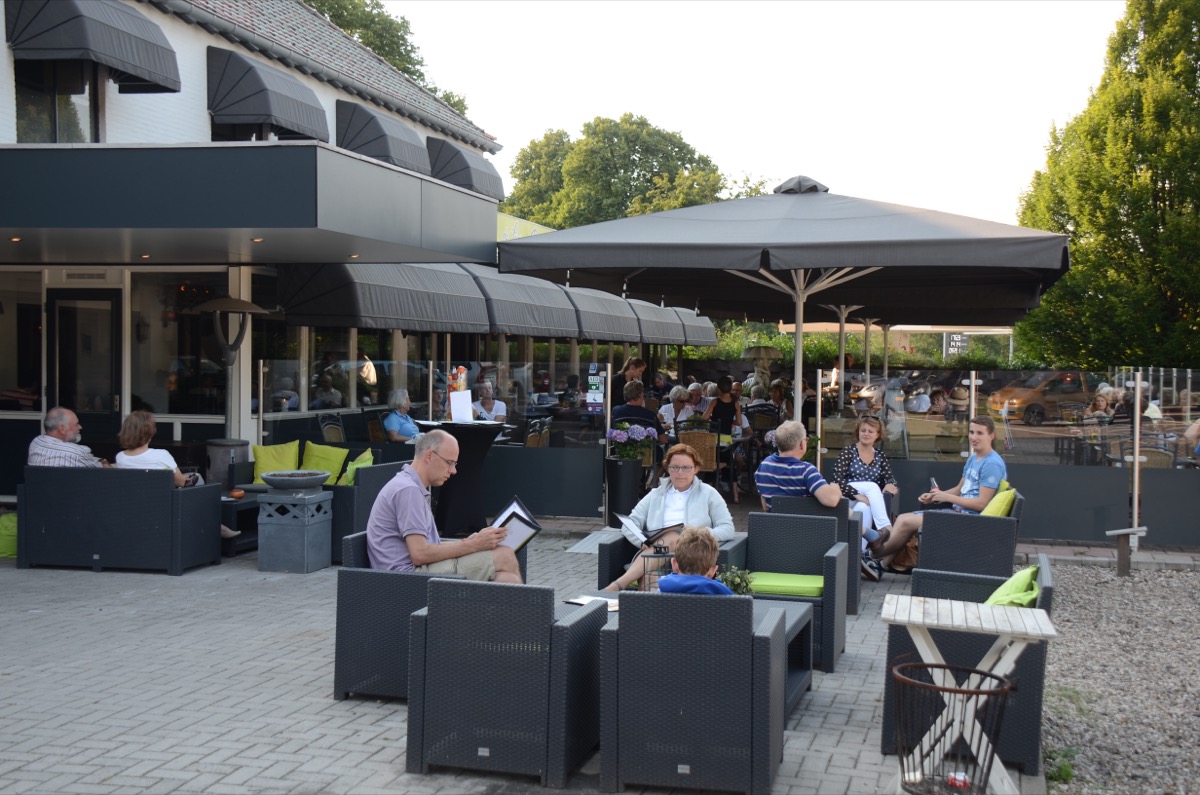 A packed outdoor restaurant terrace at dusk with several groups of guests seated at dark wicker tables and chairs under large umbrellas, some browsing menus, with more diners visible inside through the glass front.