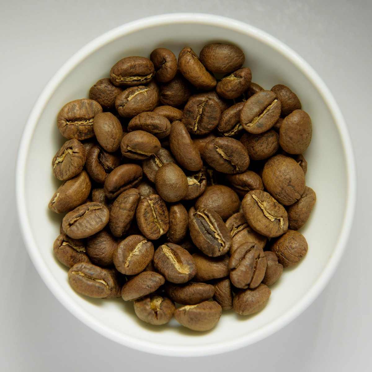 Overhead close-up of a white bowl filled with whole roasted coffee beans on a grey surface.