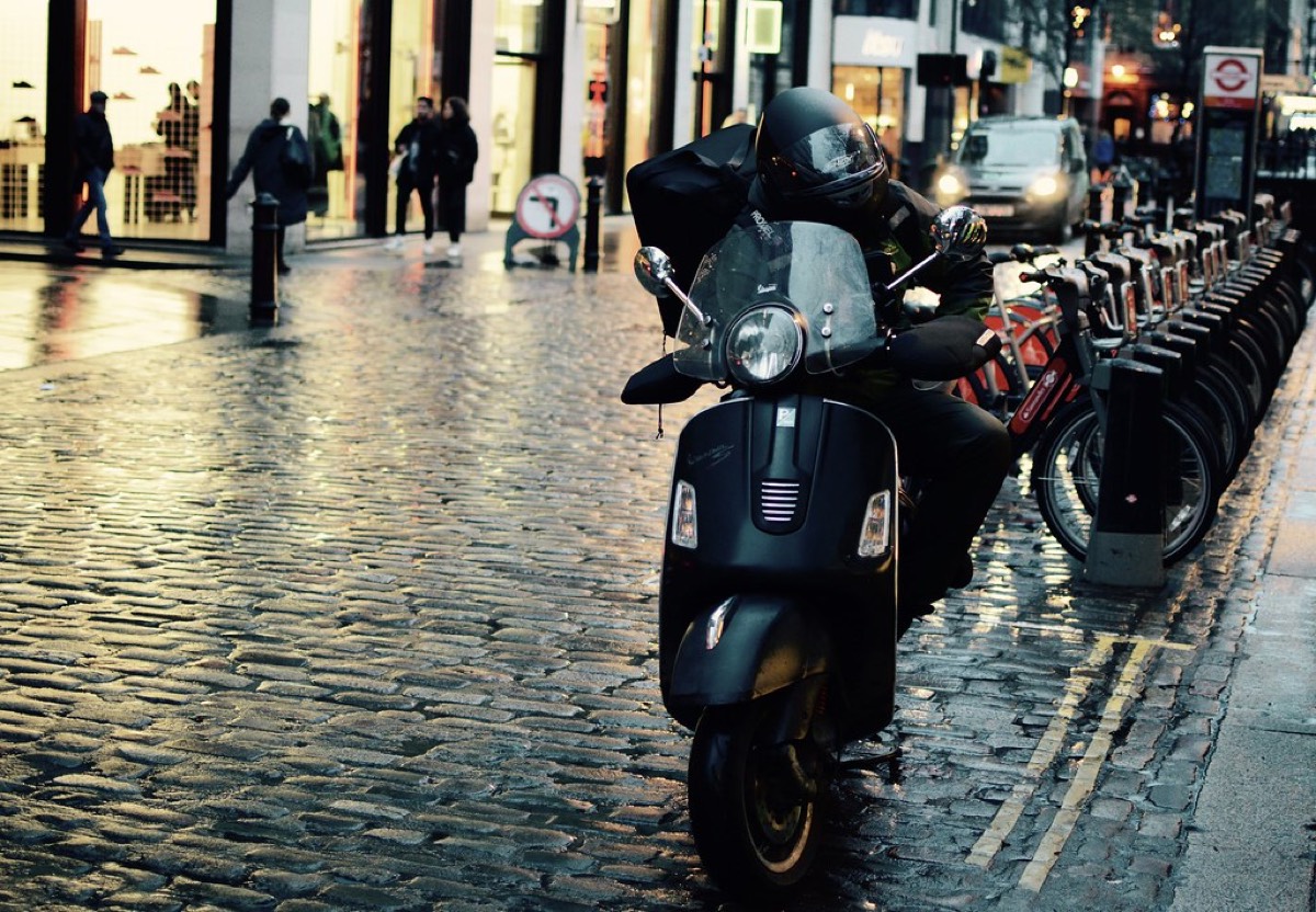 A helmeted delivery rider on a black scooter waits on a wet cobblestone city street at dusk, with a large delivery bag on their back and a row of bike-share cycles parked behind them.