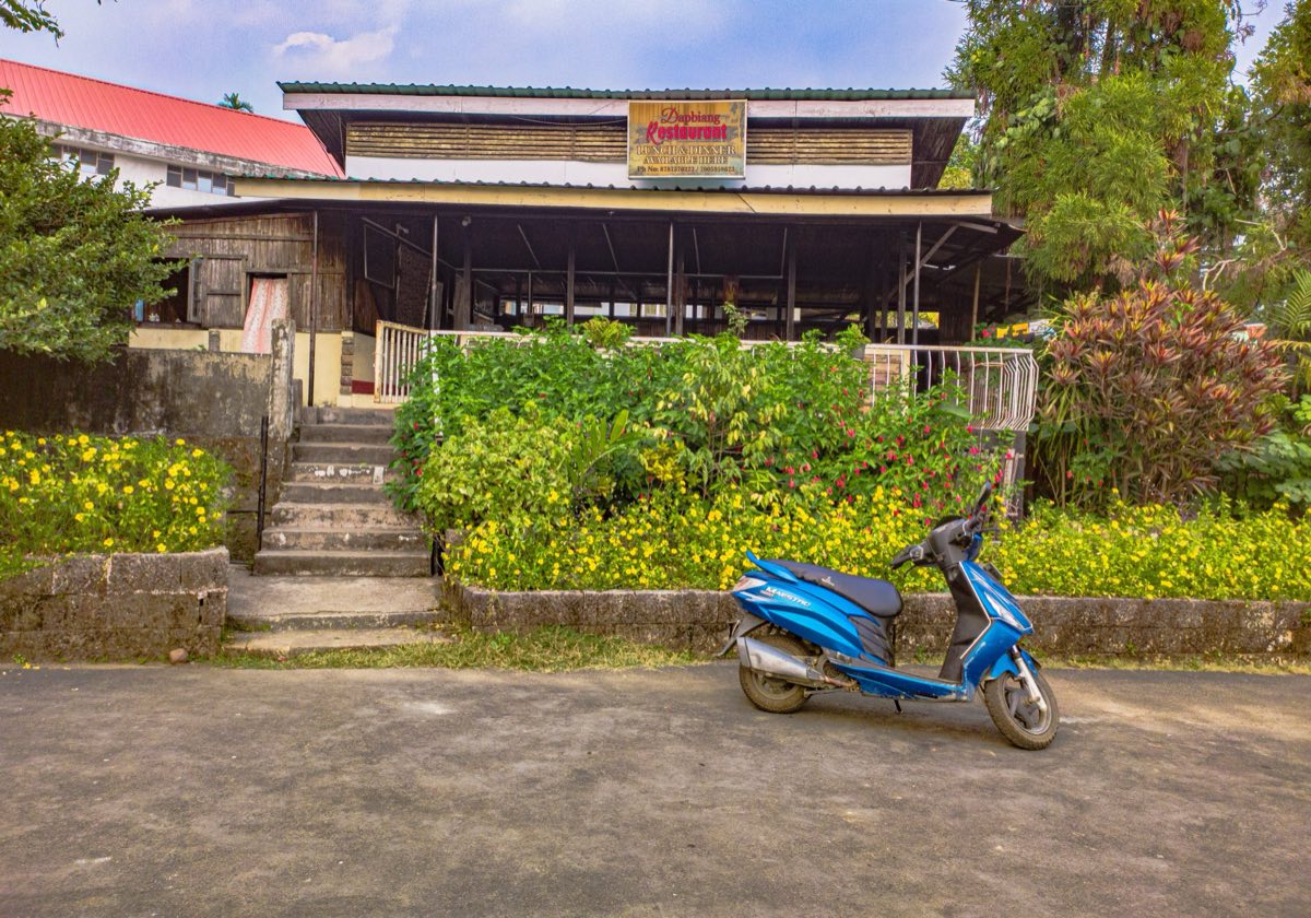 A blue scooter parked on a paved forecourt in front of a small restaurant with a wooden veranda and a sign advertising lunch and dinner.