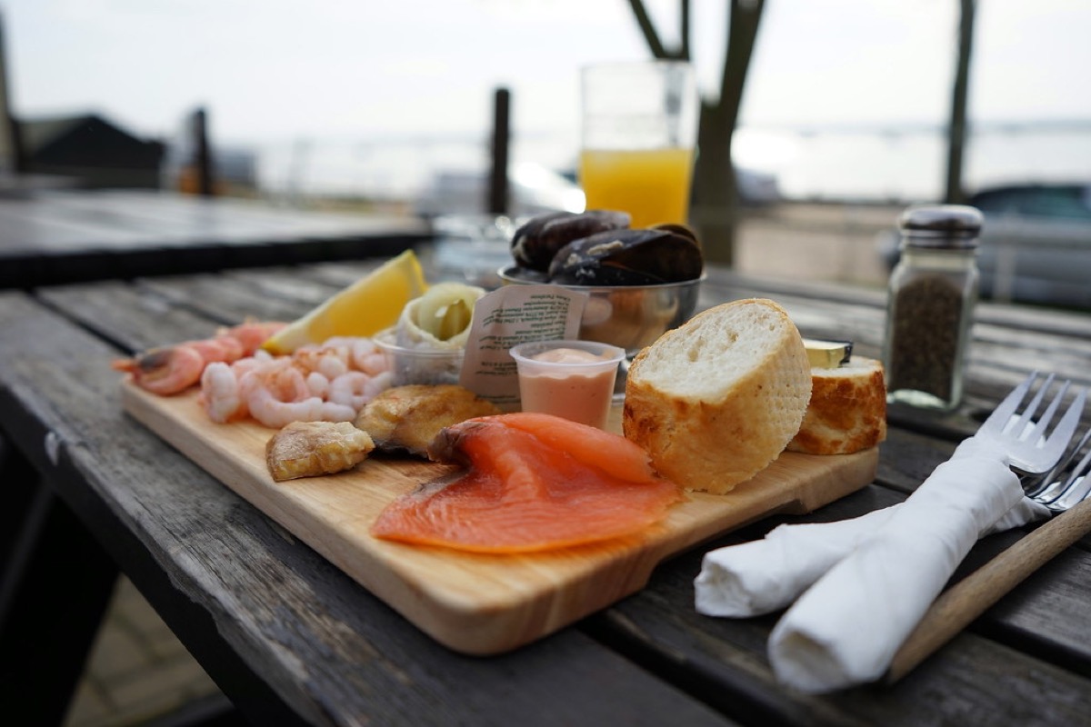 A wooden seafood board with prawns, smoked salmon, mussels, crusty bread and lemon on a table by the water