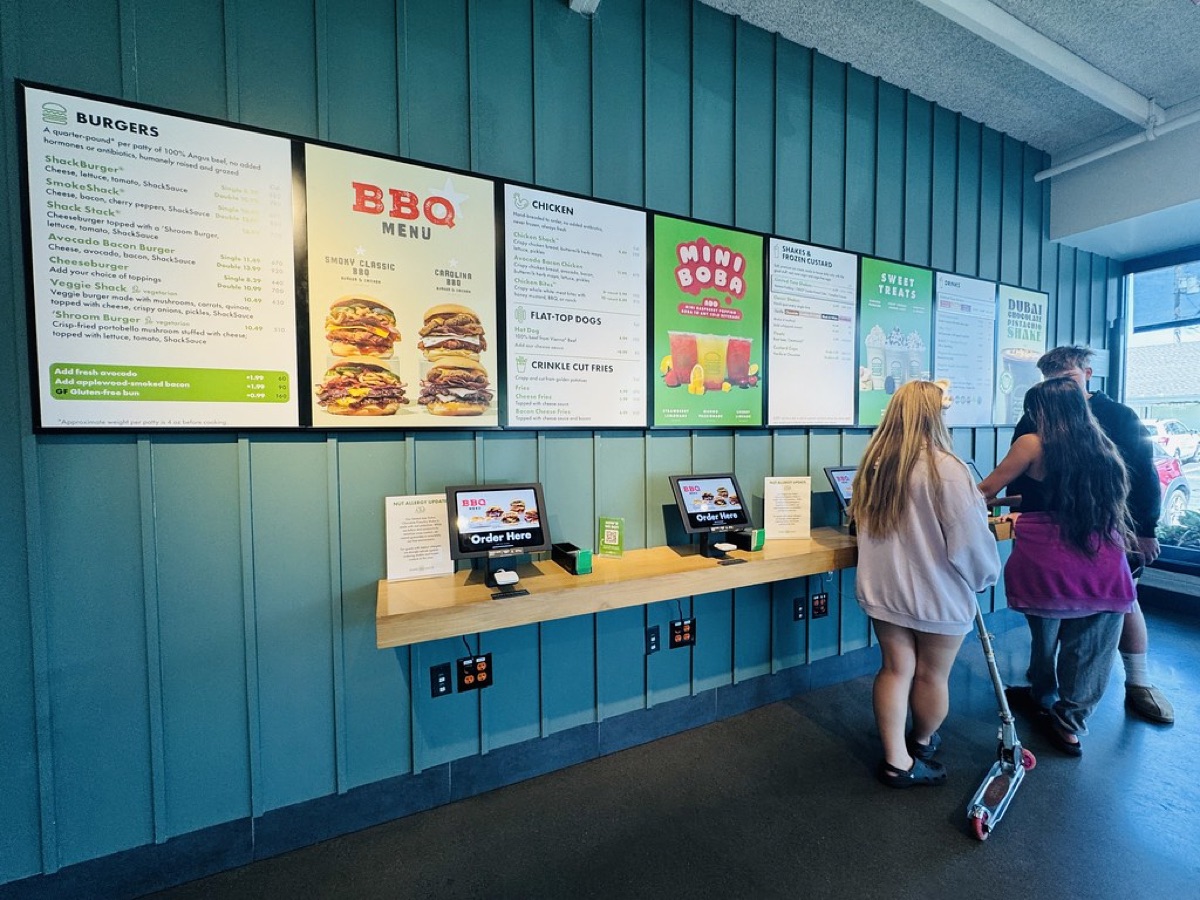 A customer orders at a row of self-ordering touchscreen kiosks below illuminated digital menu boards in a quick-service restaurant