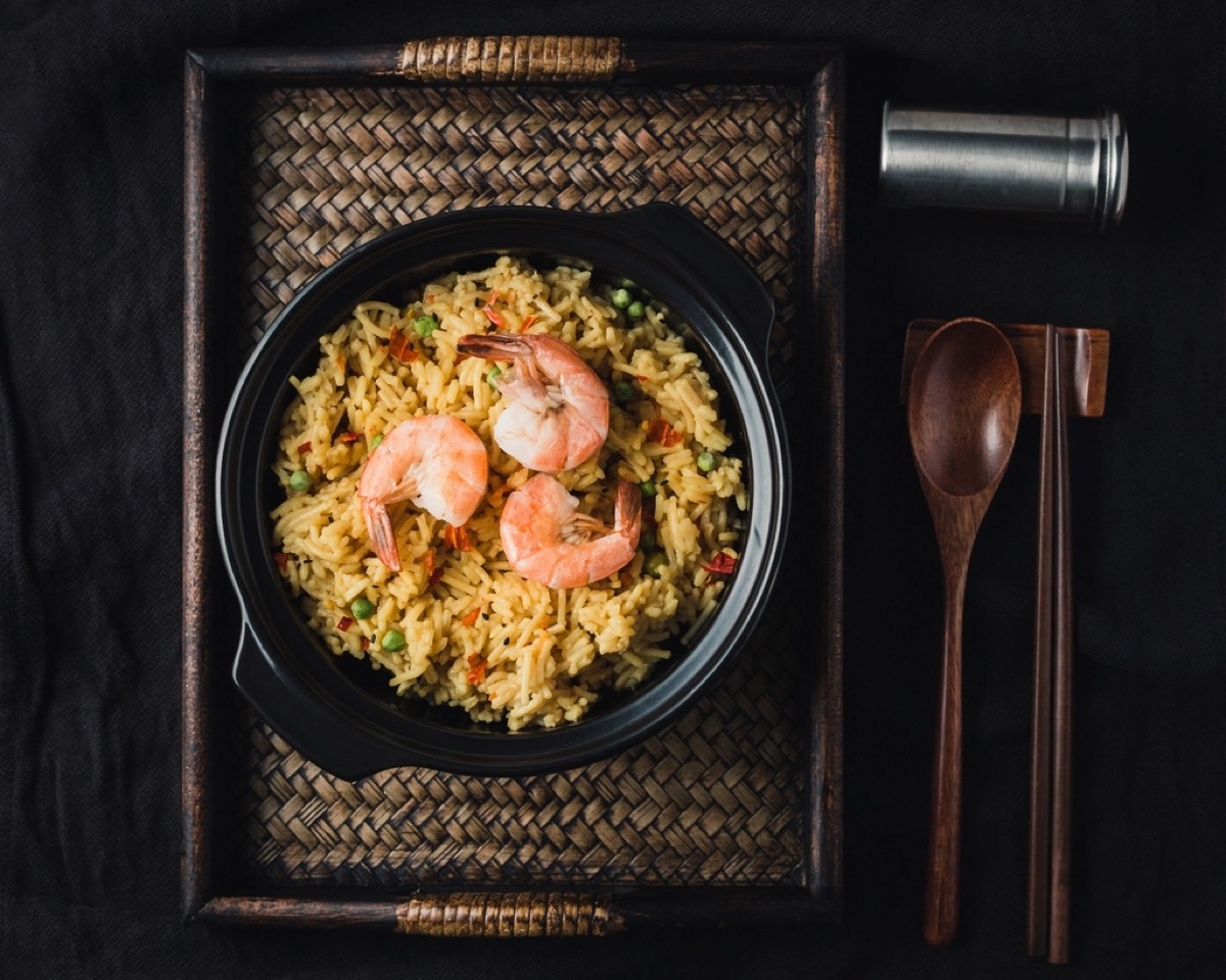 An overhead black bowl of shrimp fried rice with peas and chilli on a woven tray with a wooden spoon