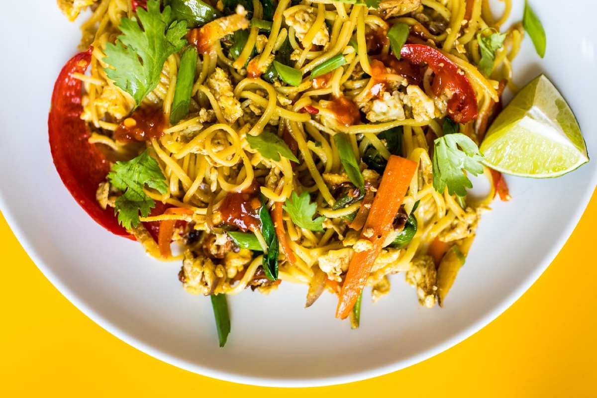 An overhead bowl of stir-fried egg noodles with carrot, red pepper, spring onion, coriander and a lime wedge
