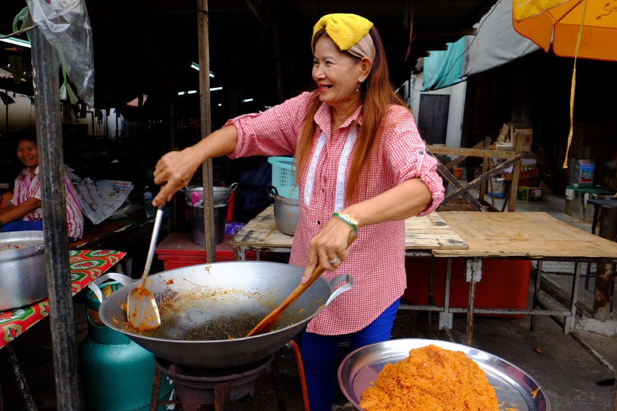 A smiling street food vendor in a checked shirt stirs noodles in a large wok over a gas burner at an open air market stall.
