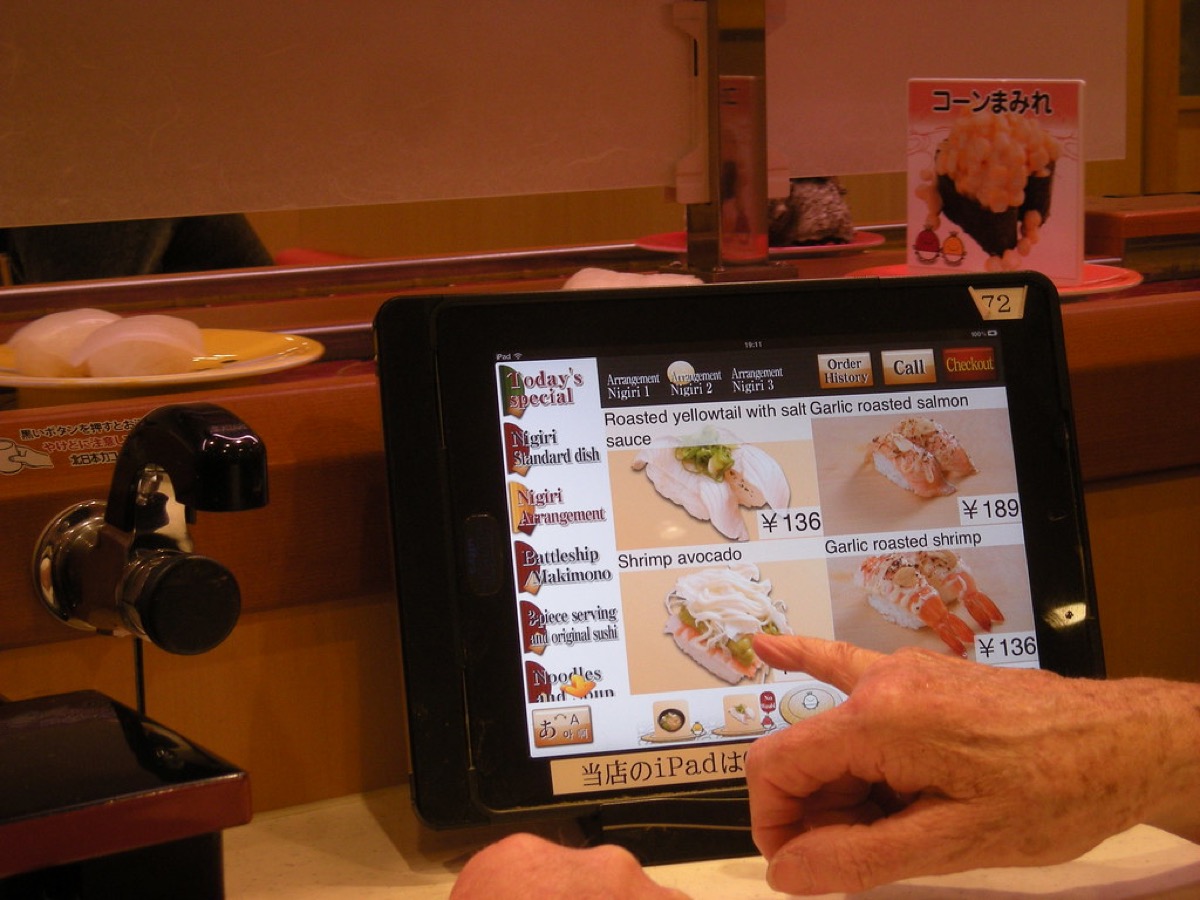 A customer's hand points at a digital menu on an iPad mounted at a restaurant counter, browsing dish photos and prices.