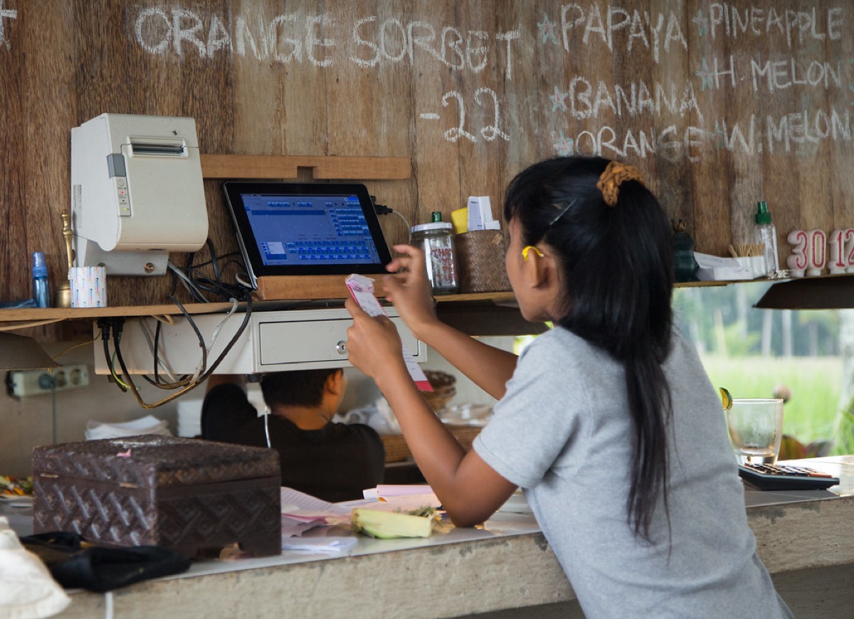 A food stall worker holds a receipt near a tablet point of sale system mounted on a wooden counter, with a cash drawer and receipt printer alongside a handwritten menu board.