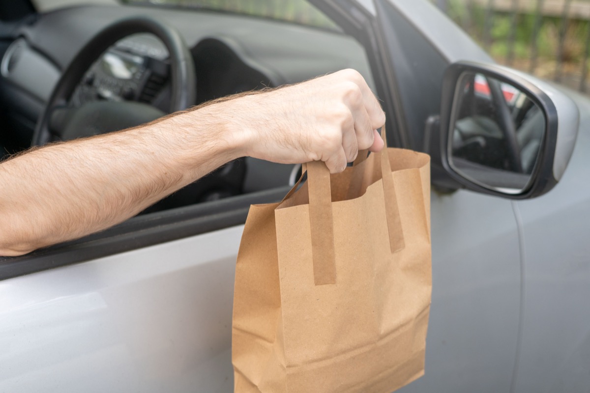 A restaurant worker's arm reaches out of a car window to hand over a plain brown kraft paper takeaway bag during a food pickup.