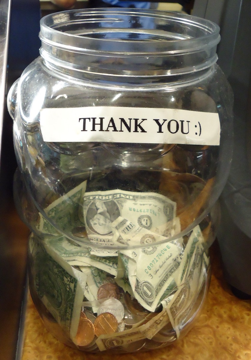 A clear tip jar labelled Thank You, filled with folded cash and coins, on a cafe counter