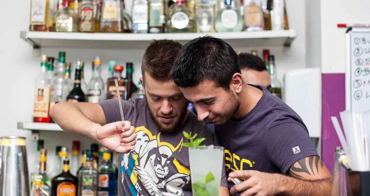 An experienced bartender closely watches and guides a trainee bartender who is building a mojito behind the bar