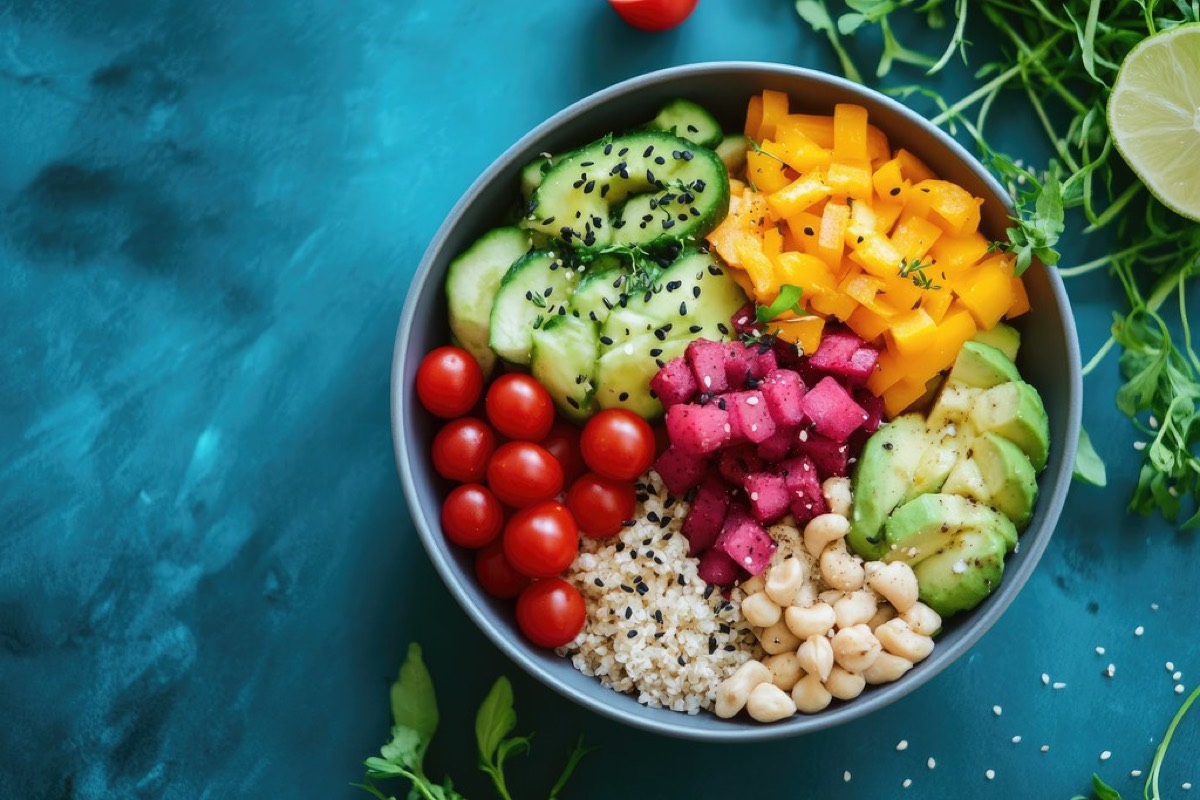 Overhead shot of a colourful vegan bowl with cucumber, cherry tomatoes, mango, watermelon radish, avocado, quinoa and cashews on a teal background