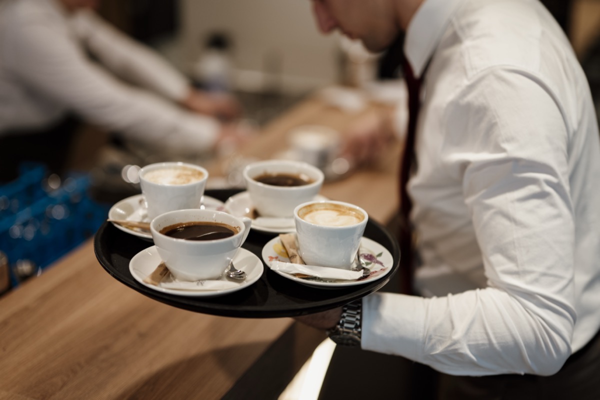 A waiter in a white shirt carries a black tray with four cups of coffee across a busy cafe counter.