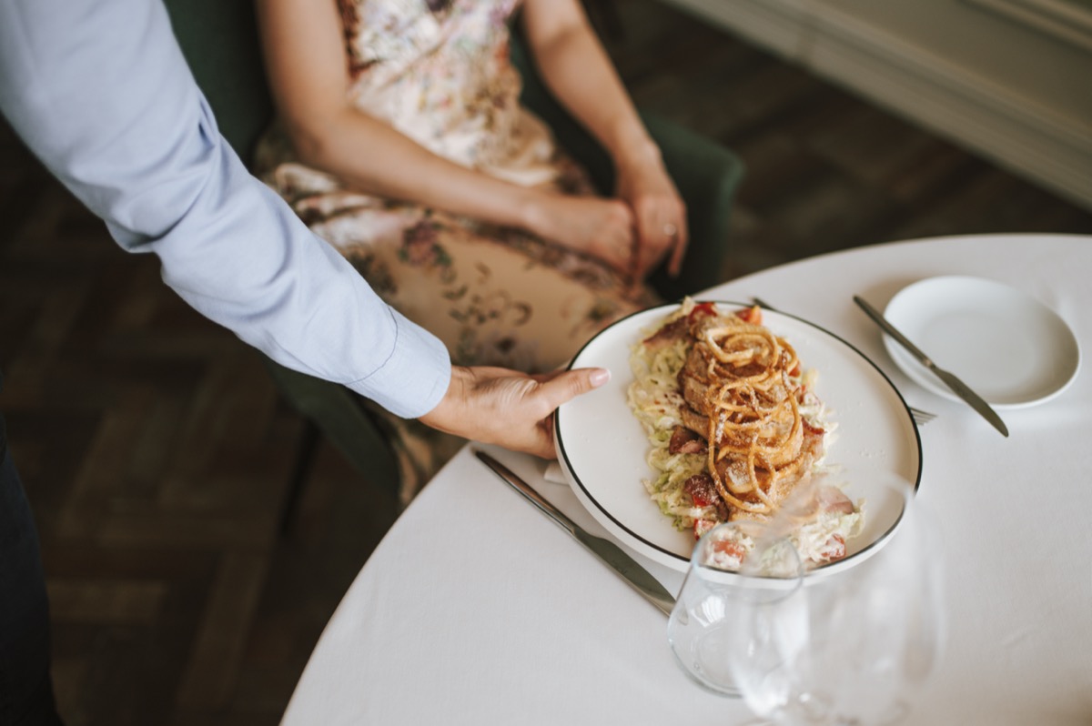 A waiter in a light blue shirt places a plated pasta dish in front of a seated guest at a white-clothed restaurant table.
