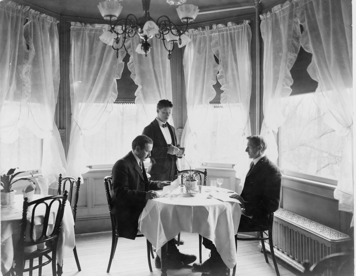 A waiter in a bow tie stands beside a dining table writing on a small notepad while two seated guests look on, in a sunlit dining room with sheer curtains.