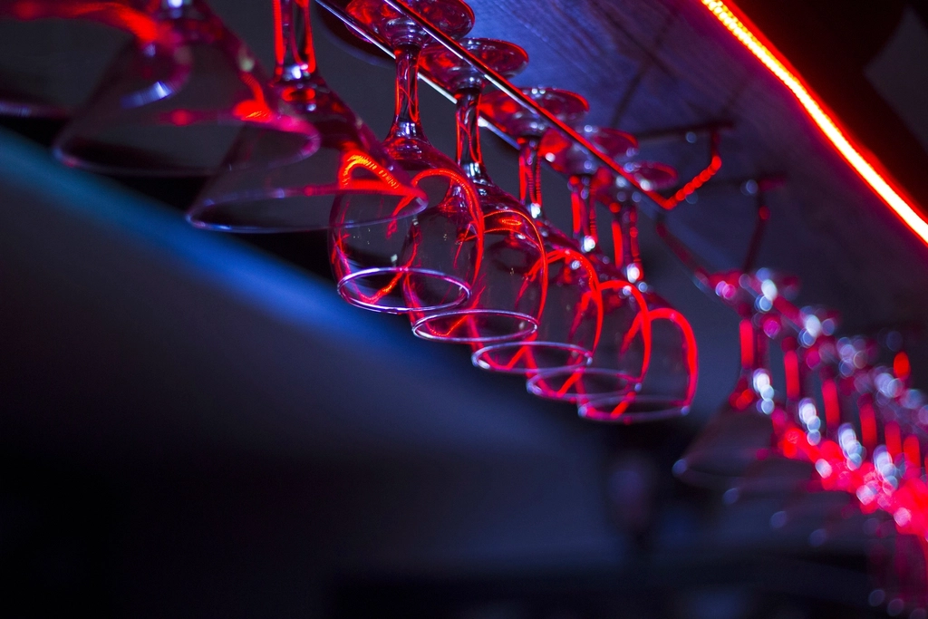 Wine glasses hanging upside down in an overhead rack above a bar, lit by red neon
