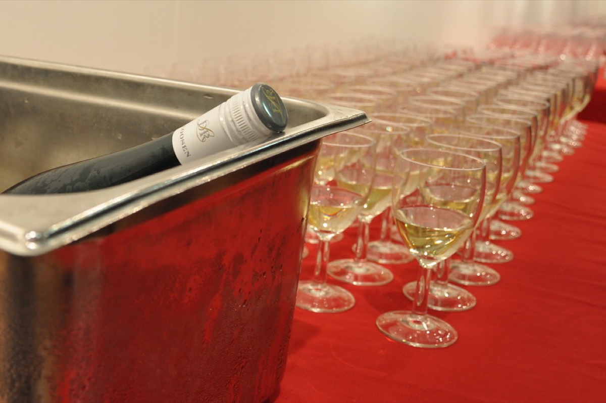A long row of wine glasses partially filled with white wine beside an ice bucket on a red-draped table