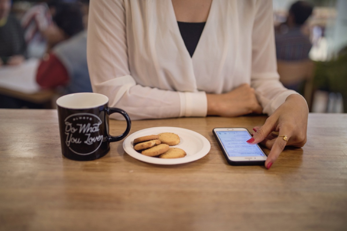 A woman seated at a cafe table with a coffee mug and a small plate of cookies, tapping the screen of her smartphone which shows an open messaging or app screen.