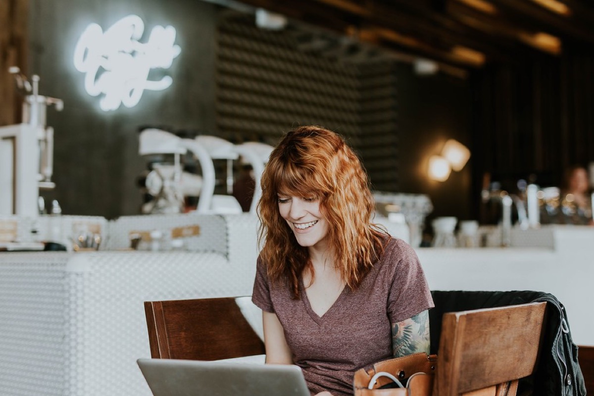 A smiling woman with curly red hair sits at a cafe table working on a laptop, with espresso machines and a neon sign blurred in the background.